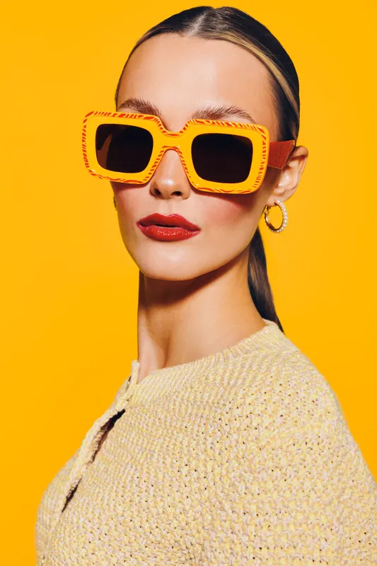 Stylish woman with slicked back hair wearing oversized yellow sunglasses and red lipstick poses against bright yellow background. Fashion concept with bold accessories and confident expression.