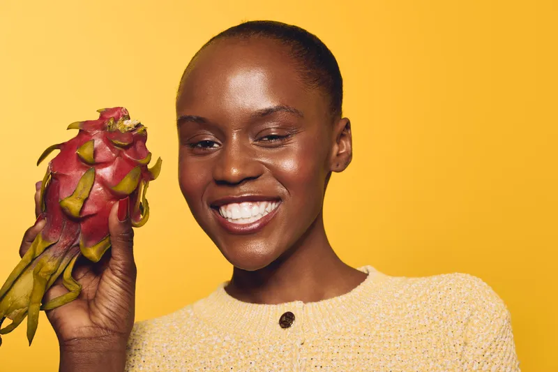 Smiling black African woman with dark skin holding a dragon fruit near her face on yellow background. Concept of healthy lifestyle, exotic fruit, and natural beauty with cheerful mood.