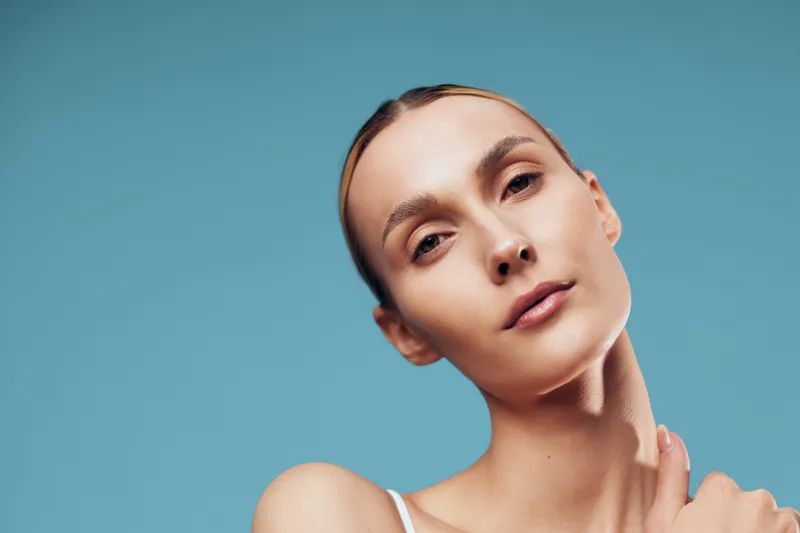 Close portrait of woman, skincare routine, hand on neck, face cream, smooth skin, soft studio light, blue background