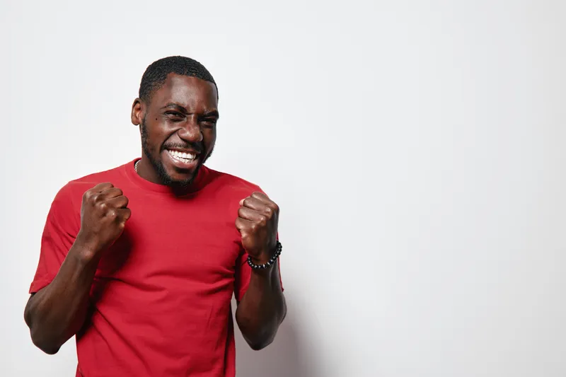African American man with dark skin wearing red t shirt celebrating success with clenched fists and joyful expression on white background. Happiness and achievement concept.