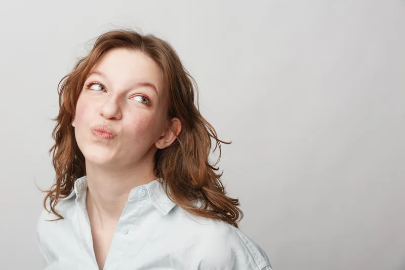 Young woman with red hair and fair skin looks thoughtfully to the side with pursed lips. Casual white shirt and neutral background create a simple portrait concept.