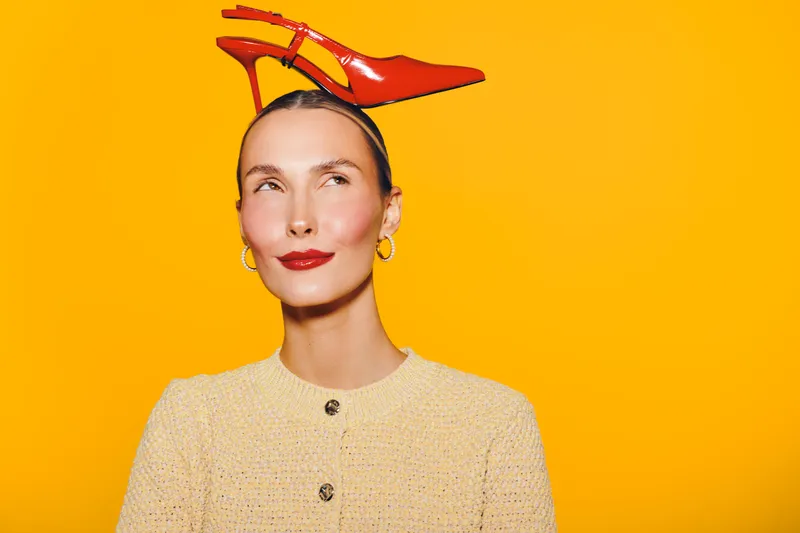 Young woman with red high heel shoes on her head looks sideways with a playful smile on bright yellow background. Concept for fashion, creativity, and fun expression.