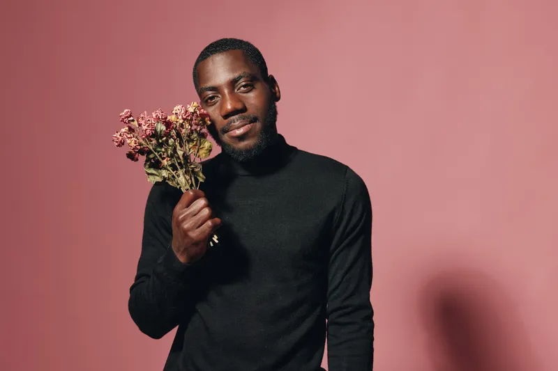 African man holding dried flowers in creative conceptual visual style with black turtleneck sweater on pink background. Portrait, smile and artistic expression for modern lifestyle and culture.