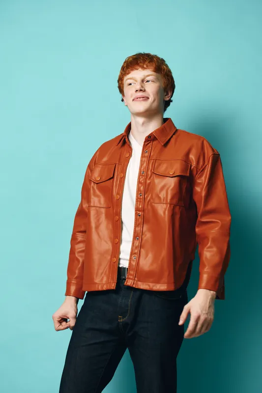 Young man with red hair wearing brown leather jacket and white shirt standing against blue background in studio. Casual style, portrait and confident pose for fashion and lifestyle.