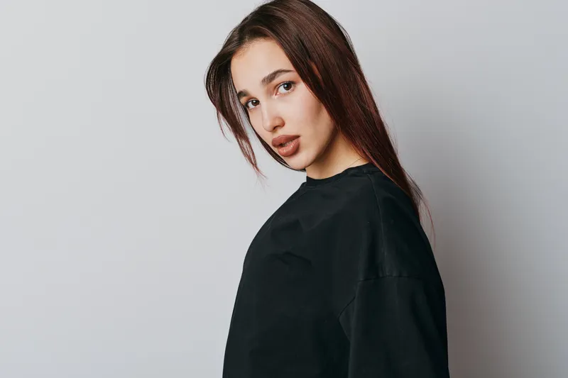 Young woman with long brown hair wearing black shirt posing against white background looking at camera with serious expression and natural makeup in studio portrait.