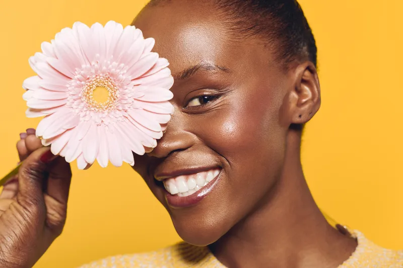 Close-up of happy African woman with dark skin holding pink flower near face smiling against yellow background. Concept of beauty, skincare, natural glow, and wellness.