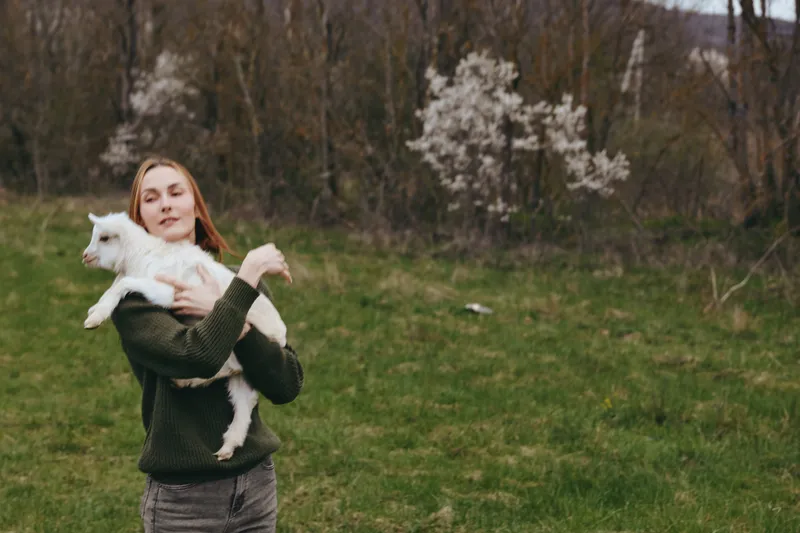 Casual woman holding a goat kid outdoors in a grassy field, warm natural light, rustic lifestyle mood, close connection with animal, joyful moment, gentle care, simple peaceful scene.