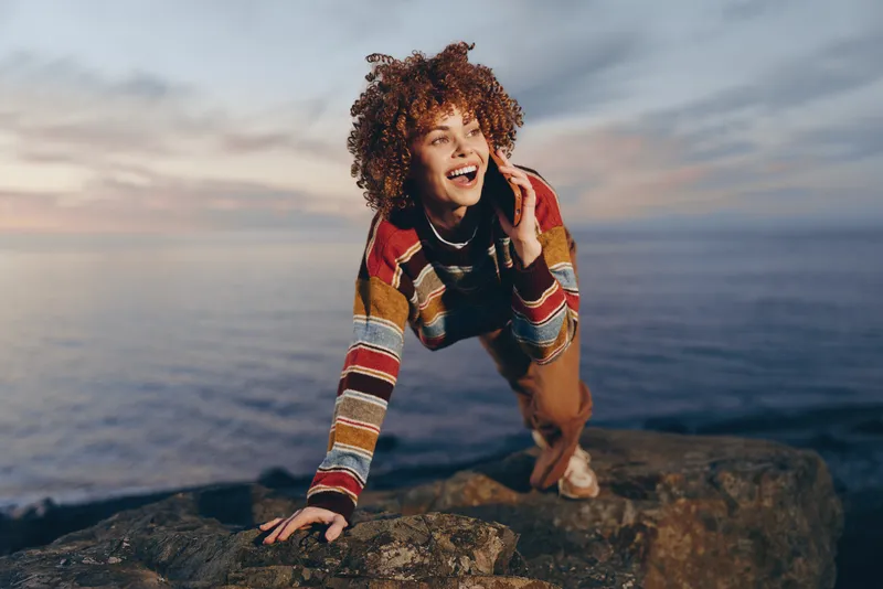 Woman lifestyle outdoors by ocean coast at sunset, curly hair wind swept, smiling and speaking on phone while standing on rocky cliff, casual travel mood, cinematic natural light