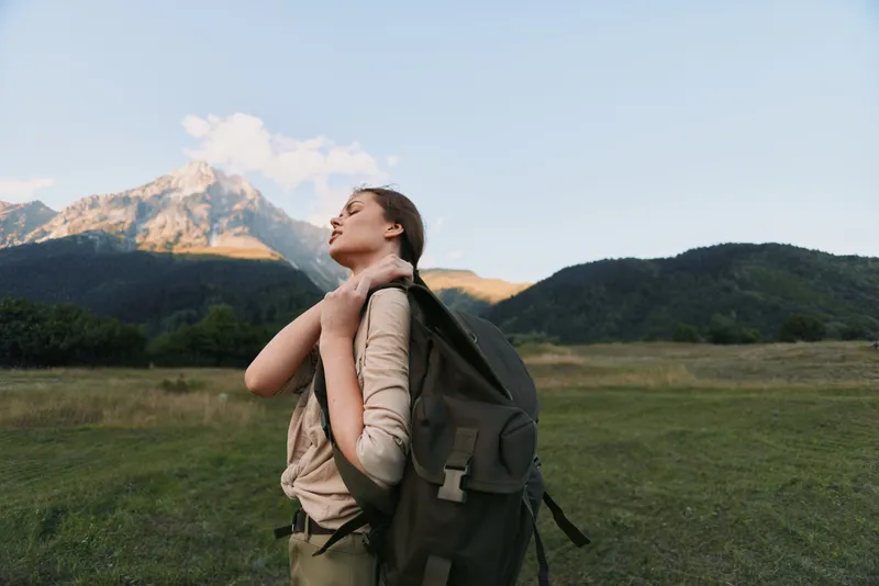 Young woman with long hair enjoys a peaceful moment outdoors with a backpack in a mountain meadow during sunset, expressing relaxation and freedom.