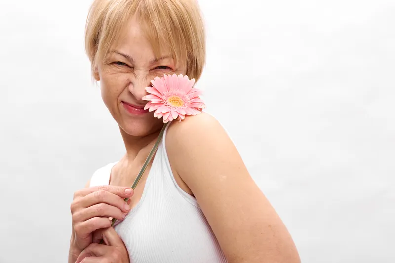 Smiling woman holding pink flower near lips, closeup portrait against white background, spring freshness, soft studio lighting, calm joyful expression
