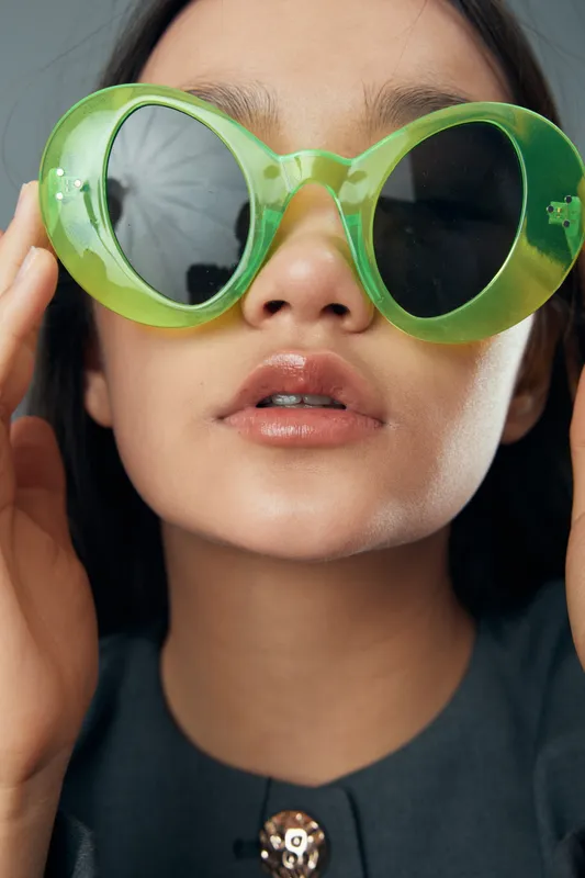 Woman wearing large green sunglasses with round lenses touching the frames closeup portrait with neutral lips and smooth skin on light background in fashion style.