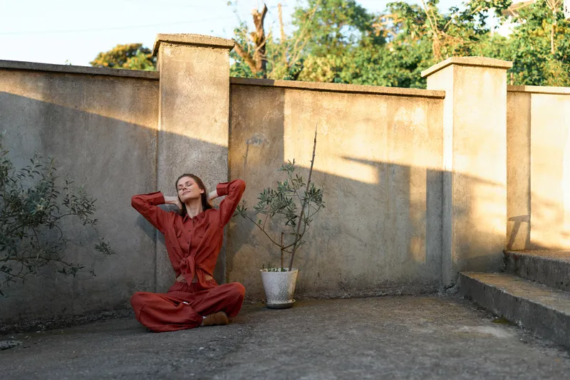 A woman in a red outfit sits on a concrete courtyard wall, stretching her arms behind her head with a calm, meditative mood under bright outdoor light