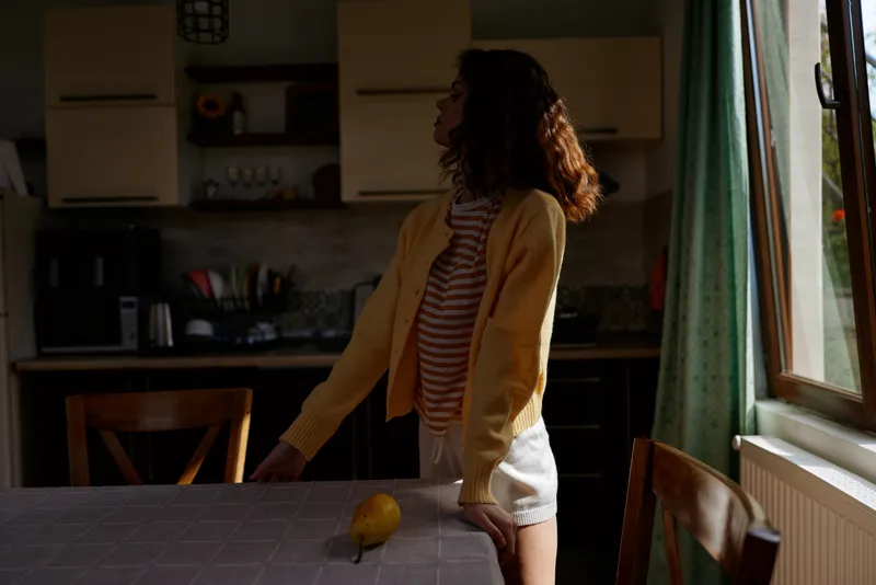Woman in a cozy home kitchen stands beside a table with a lemon, wearing a striped shirt and yellow cardigan, enjoying a calm morning moment in natural light