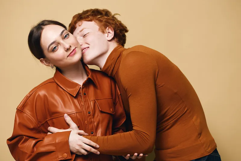 Couple with affectionate embrace and tender kiss on cheek, woman in leather jacket and man in brown sweater posing together in studio with beige background and warm mood.