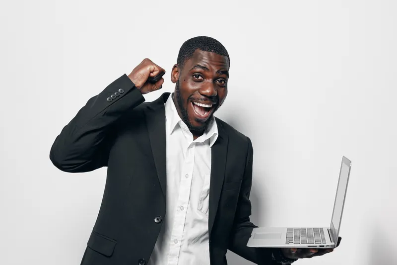 African American man with dark skin wearing black suit and white shirt holding laptop and celebrating success with fist pump and happy expression on white background.