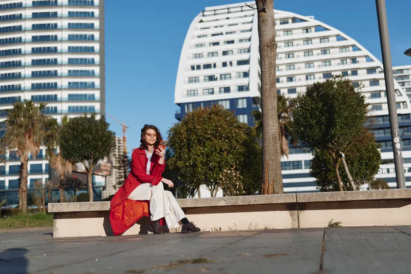 Young woman wearing a red coat sits on a concrete bench in an urban park on a sunny day, enjoying a relaxed moment with a bright blue sky behind modern buildings.