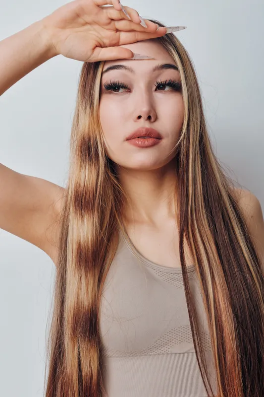 Young woman with long straight hair posing with hand on forehead looking thoughtful and relaxed against plain light background. Closeup portrait with natural makeup and neutral expression.