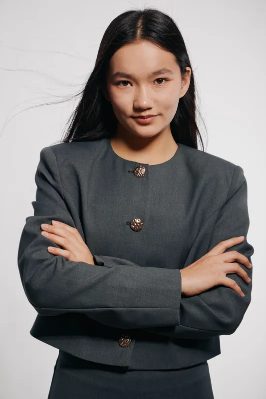 Young confident woman with long black hair and gray business jacket standing with crossed arms looking at camera serious expression. Studio portrait with plain background.