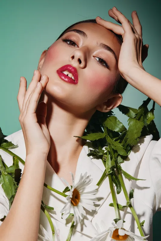 Close up of fashion model with artistic floral accessories posing against a green background. She wears bold makeup and glossy lips expressing beauty creativity and fashion concept.
