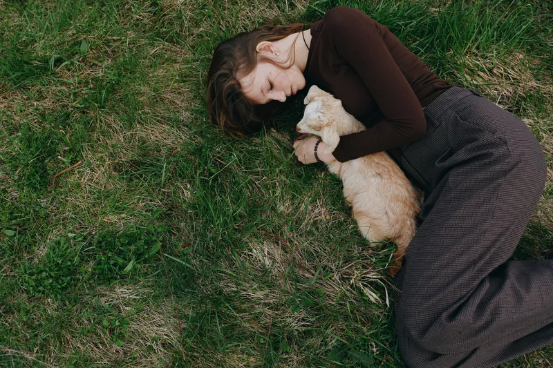 Young woman lies on green grass outdoors embracing a small baby goat in a tender moment of connection and relaxation in casual autumn clothes.