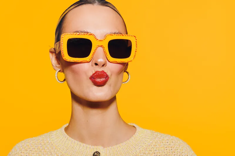 Young woman with bright orange sunglasses and red lipstick makes a kiss face on vibrant yellow background. Fashion portrait with trendy accessories and bold colors.