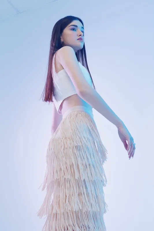 Young woman in white sleeveless top and fringed skirt poses confidently in studio with soft blue lighting. Concept of woman beauty fashion and modern style.