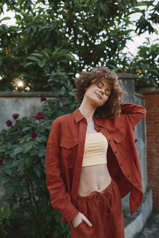 A confident woman poses outdoors in casual fashion, wearing a rust jacket and crop top in a garden setting. Bright light highlights a relaxed lifestyle and style