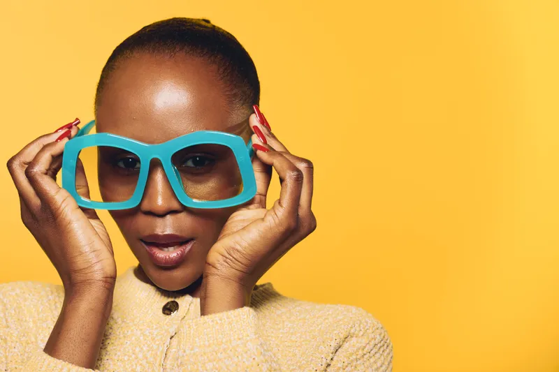 Portrait of confident dark skinned African woman wearing large blue sunglasses. She poses against yellow background in beige sweater. Concept for fashion, style, and beauty with vibrant colors.