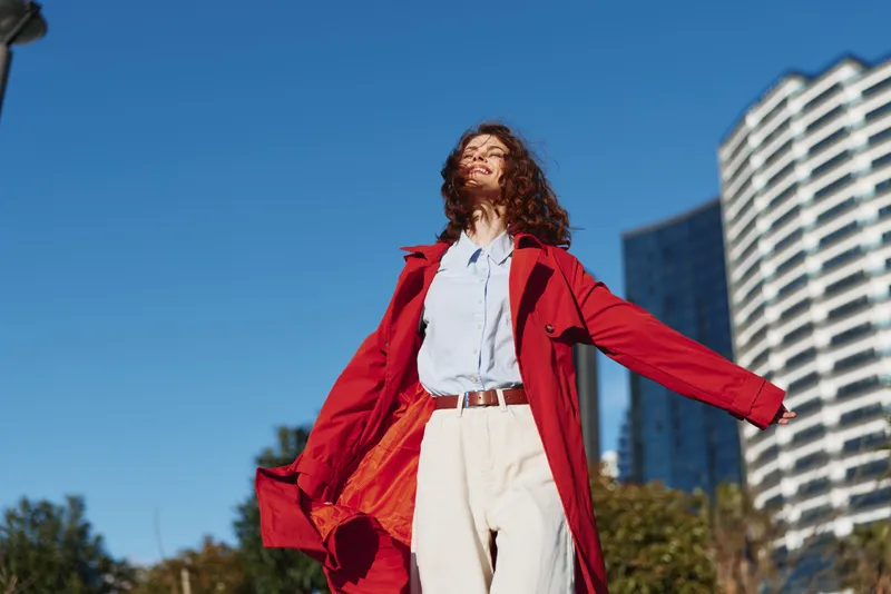 A woman in a red coat stands outdoors with arms outstretched under a bright blue sky, white blouse and light trousers, embodying carefree confidence and urban freedom.