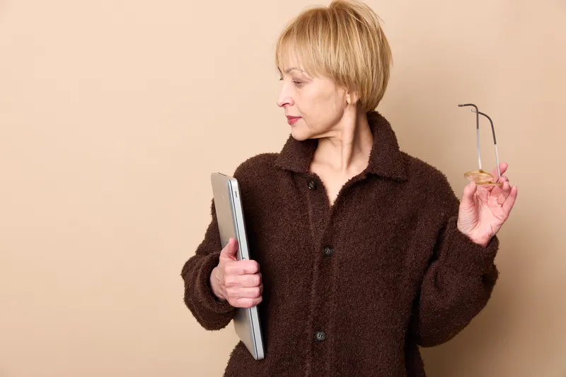 Senior woman indoors holding smartphone and glasses, wearing a brown coat against a beige wall, focused expression, casual lifestyle, soft studio light