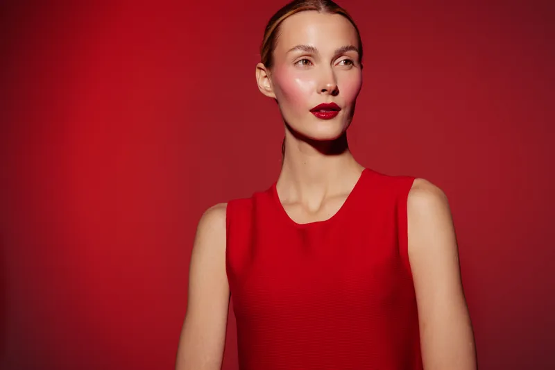 Portrait of a confident woman in a red sleeveless top against a red background. She has a sleek hairstyle and bold red lipstick. Concept of fashion, beauty, and elegance.