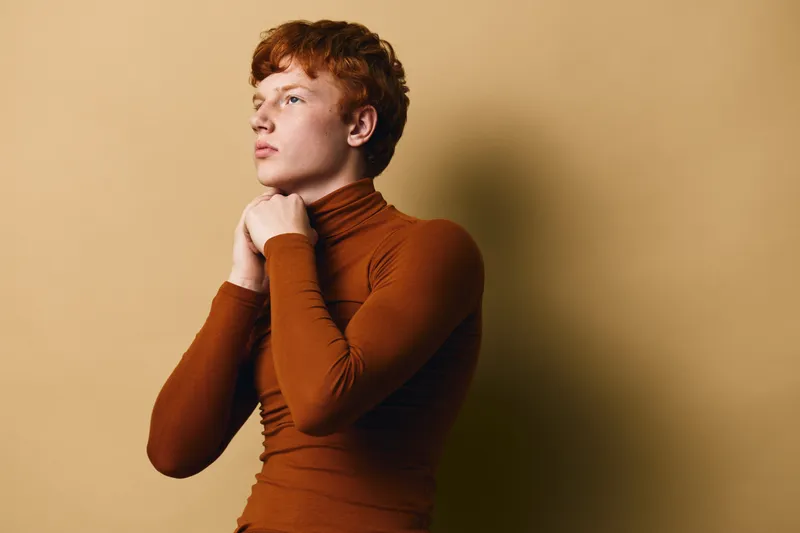 Young man with red hair in brown turtleneck sweater posing against beige background for fashion portrait and casual style with thoughtful expression and profile view.
