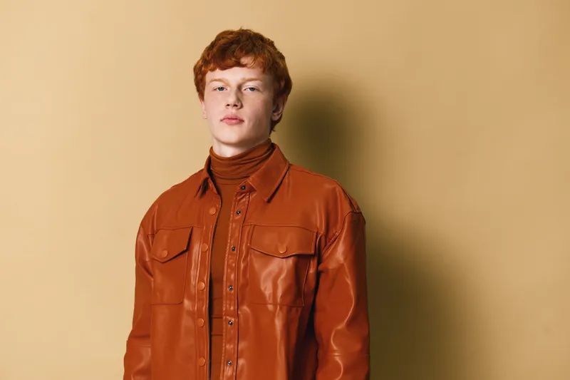 Young man with red hair wearing brown leather jacket and turtleneck standing against beige background looking confident and serious with relaxed posture in studio.