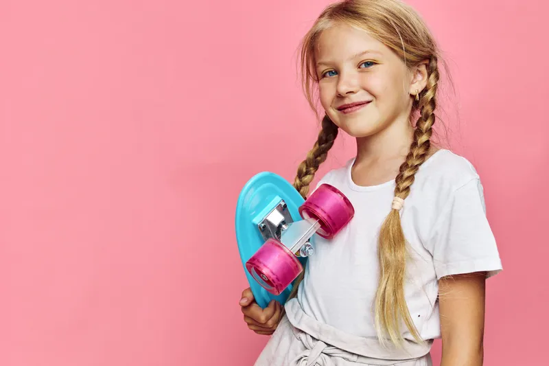 Smiling girl with blonde braids holds a blue skateboard with pink wheels against a pink background. Casual outfit and confident expression in studio lighting.