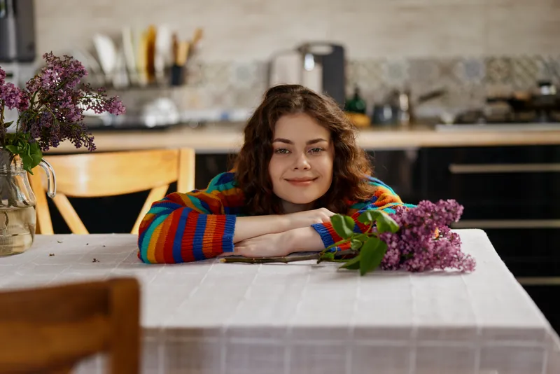 A woman in a colorful striped sweater rests her chin on folded arms over a white table in a bright home kitchen, conveying a cozy lifestyle moment.