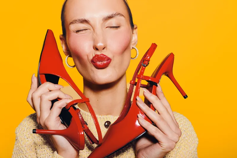 Happy woman with closed eyes holds red high heel shoes close to face on bright orange background. Concept of fashion, style, footwear, and joyful emotions in studio lighting.