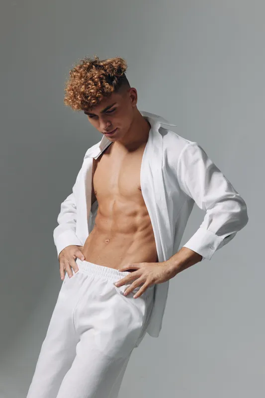 Young man with curly hair wearing white pants and open shirt showing toned abs and muscular body posing against gray background in studio with soft lighting.