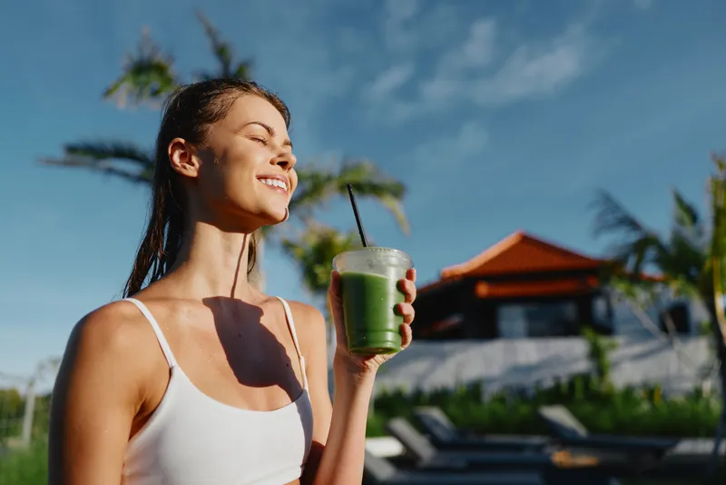 A healthy lifestyle outdoors woman enjoys a smoothie in sunny resort setting, smiling while holding a green drink with a straw; a poolside scene conveys wellness, relaxation, and a leisurely summer