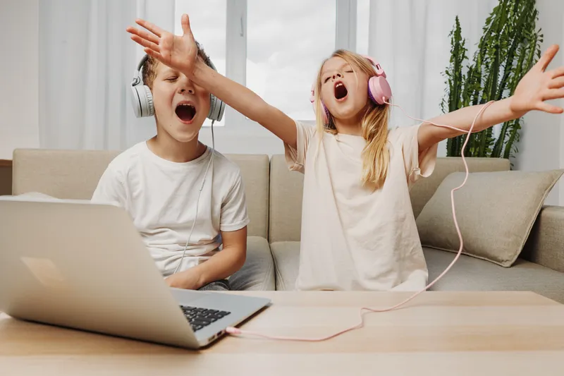 Two excited children wearing headphones sing loudly with open mouths while sitting on a couch in a cozy living room. Concept of childhood joy, music fun, and family entertainment.