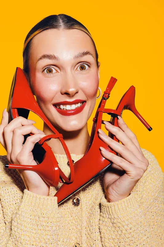 Happy woman with red lipstick holds red high heel shoes near face on yellow background. Concept of fashion, style, and joyful emotions in studio portrait photography.