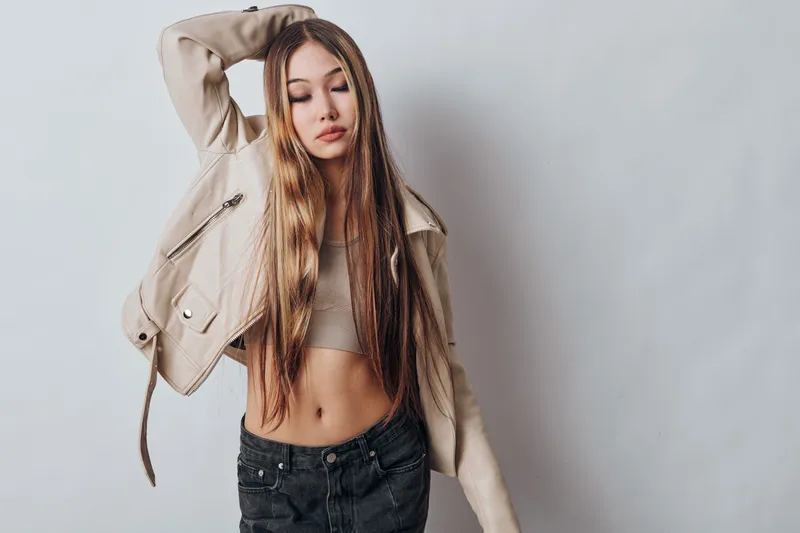 Young woman with long hair wearing beige jacket and crop top standing against gray background with closed eyes and relaxed expression for fashion portrait.
