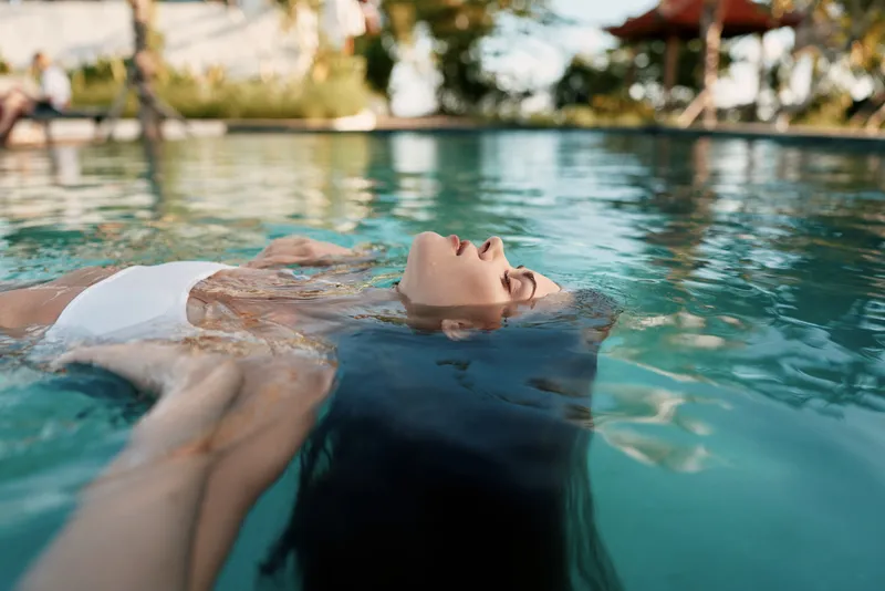 A woman floats serenely in a clear pool, enjoying relaxation under bright sunlight, a lifestyle moment of leisure and escape, with calm water, reflective surface and poolside tranquility in a sunny