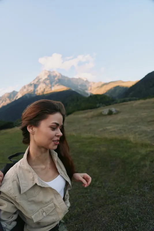 Woman in casual jacket posing outdoors on a mountain hillside, enjoying fresh air and expansive scenery, lifestyle portrait with natural light, distant peaks, and tranquil mood.