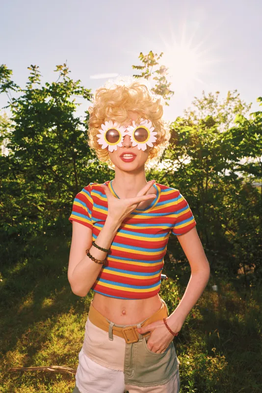 Playful woman wearing a striped crop top and daisy sunglasses poses outdoors in warm sunlight, embracing retro film color, casual summer lifestyle, carefree and vibrant, with lush greenery behind.
