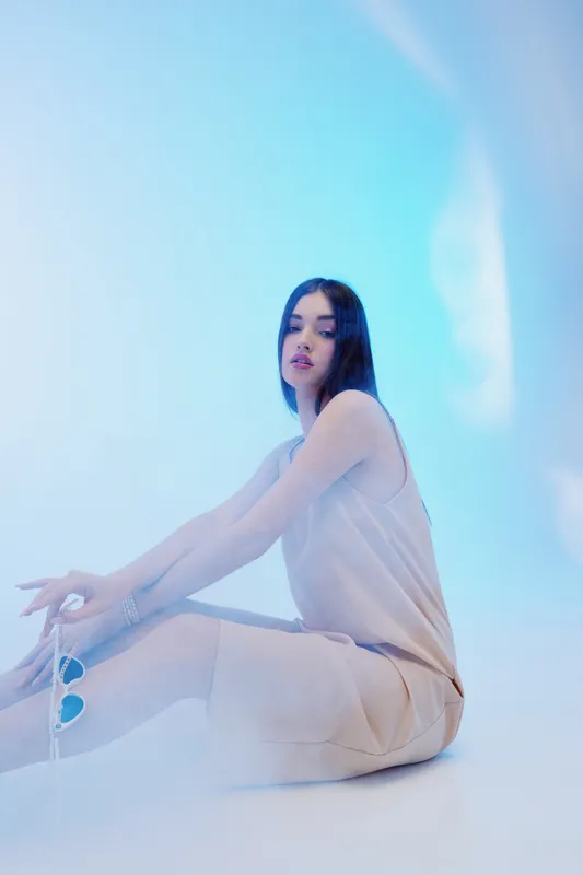 Young woman beauty fashion model sitting on the floor in a soft beige dress against a light blue background. Concept of elegance, style, and modern female fashion portrait.