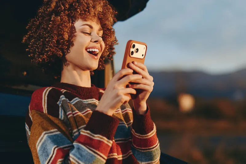 Cheerful woman outdoors, laughing with a smartphone, curly hair and plaid sweater in warm sunset light, relaxed lifestyle, soft breeze, countryside background golden evening atmosphere today