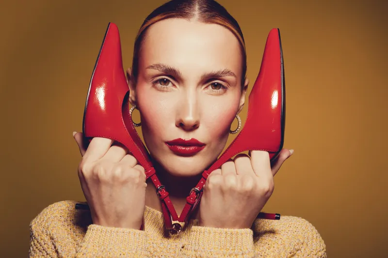 Portrait of a woman holding red high heel shoes near her face against a warm brown background. Concept of fashion, style, elegance, and beauty with bold makeup and accessories.