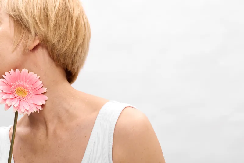 Blonde woman closeup of neck and shoulder with a pink flower on soft skin, minimal bright white studio background, beauty and skincare concept for gentle freshness and calm care