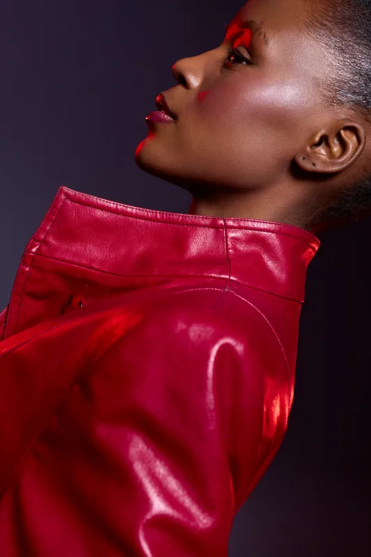 Profile of a dark skinned African woman wearing a red leather jacket with glossy makeup and red eyeshadow. Studio portrait with dramatic lighting and fashion concept.