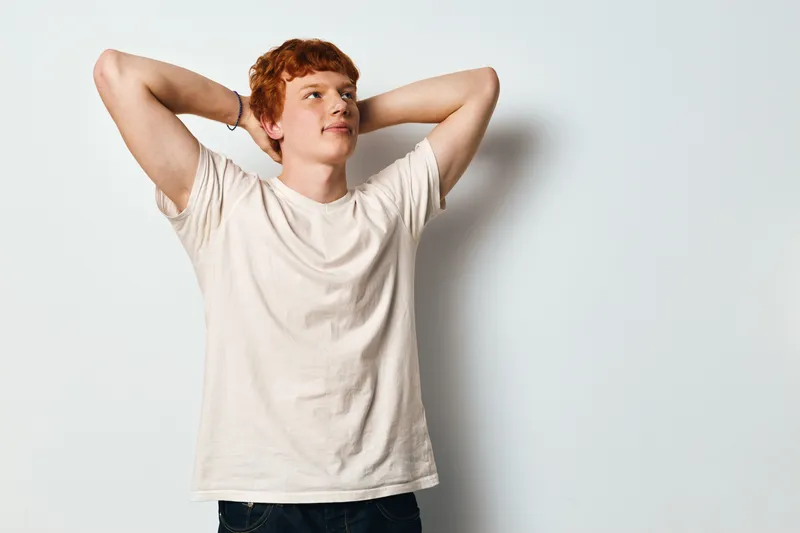 Young man with red hair in casual shirt standing relaxed with hands behind head looking away on white background. Portrait of thoughtful expression and confident posture.